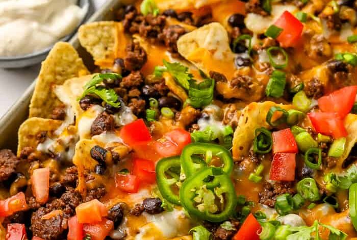 A tray full of baked nachos topped with ground beef, black beans, green onions, tomatoes, jalapenos and shredded cheese, with a side of salsa and sour cream